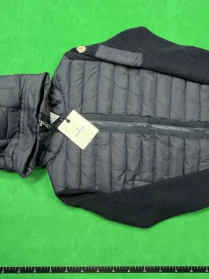 Moncler cardigan  - Stilren svart dunväst från Moncler med stickade sidor och rygg, quiltad framsida och en praktisk huva. Västen har dragkedja framtill och Moncler-logga på axeln. Perfekt för lager-på-lager och snygg till streetwear. Passar För både S och m