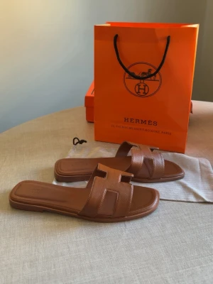 Bruna Oran sandaler i läder - Snygga bruna Oran sandaler i läder med den ikoniska H-designen över foten. Platta sulor och öppen tå gör dem perfekta för sommaren. Klassisk och stilren modell som är lätt att matcha med olika outfits.