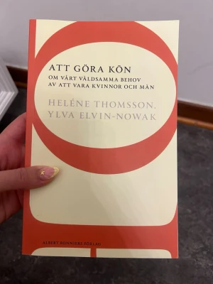 Att göra kön av Thomsson & Elvin-Nowak - Boken 'Att göra kön' av Heléne Thomsson och Ylva Elvin-Nowak utforskar könsroller och samhällets syn på kvinnor och män. Omslaget är beige med stora röda cirklar och text i svart och guld. Perfekt för dig som gillar genusfrågor och samhällsdebatt.