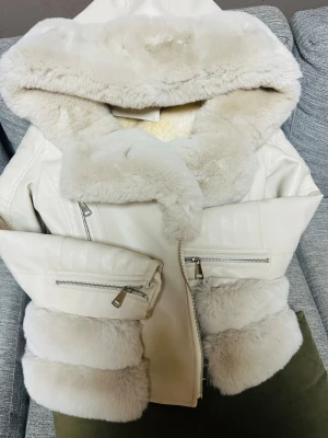 Vit pälsjacka med huva och dragkedjor - Snygg vit pälsjacka med stor fluffig huva och breda pälsdetaljer på ärmar och nederkant. Jackan har två snedställda dragkedjefickor framtill och stängs med dragkedja. Perfekt för kalla vinterdagar och ger en lyxig känsla.