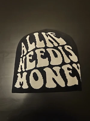 Svart mössa med vit text - Cool svart mössa med stor vit text framtill som säger 'ALL HE NEED IS MONEY' i retroinspirerad font. Mössan är mjuk och stretchig, perfekt för dig som vill sticka ut med din stil. Materialet känns som bomull eller bomullsblandning.