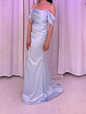 Ljusblå festklänning/balklänning - Elegant ljusblå långklänning i satin med offshoulder-ärmar och draperad midja. Klänningen har en slits på sidan och en figurnära passform som ger en lyxig känsla. Perfekt för speciella tillfällen.