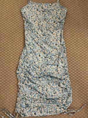 Blommig blå kortklänning med snörning - Söt blå kortklänning med små vita och gula blommor. Klänningen har smala axelband, figurnära passform och snörningsdetaljer på sidorna. Perfekt för varma dagar och ger en fräsch sommarkänsla.