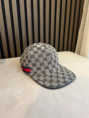 Keps GG Mörkblå biege  - Snygg keps från Gucci i beige canvas med klassiskt GG-monogram över hela kepsen. Röd och blå rand på sidan ger en extra touch. Justerbar rem baktill i svart läder. Perfekt accessoar för en trendig look.