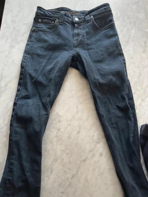 Mörkblå slim fit jeans från Nudie - Snygga mörkblå slim fit jeans från Nudie Jeans med klassisk femficksdesign och subtila slitningar. Jeansen har raka ben, normal midja och är tillverkade i mjuk denim med stretch för extra komfort. Perfekta för en avslappnad och trendig look.