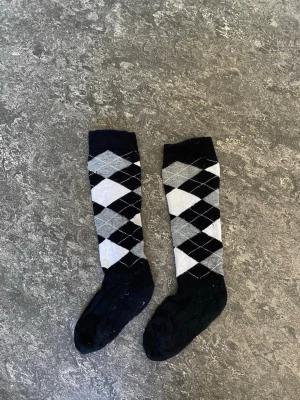 Svarta och grå rutiga knästrumpor - Säljer ett par klassiska knästrumpor med rutigt argyle-mönster i svart, grått och vitt. Strumporna är tillverkade i mjuk bomullsmix och passar perfekt för dig som vill ha en stilren och tidlös look. Märkning 'high quality' på insidan.