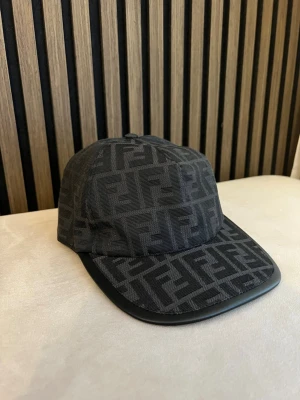 Svart keps från F med FF-mönster - Snygg svart keps från Fendi med det klassiska FF-monogrammet över hela kepsen. Kepsen har böjd skärm med läderdetalj och justerbar baksida. Perfekt för dig som gillar exklusiva accessoarer och streetstyle.