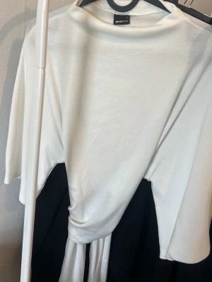 Vit omlottopp från Gina Tricot - Vit omlottopp från Gina Tricot med trekvartsärm och lös passform. Toppen har en snygg knytning framtill och är tillverkad i ett mjukt, tunt material som känns skönt mot huden. Perfekt att styla med jeans eller kjol.
