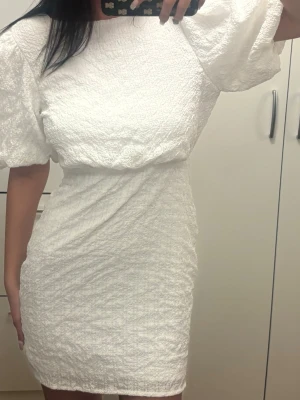 Vit kort klänning med puffärm och rosett Bubbleroom - Superfin vit kort klänning med puffärmar och snygg rosett i ryggen. Klänningen har en strukturerad yta och markerad midja som ger en smickrande siluett. Perfekt för student eller sommarens tillfällen!