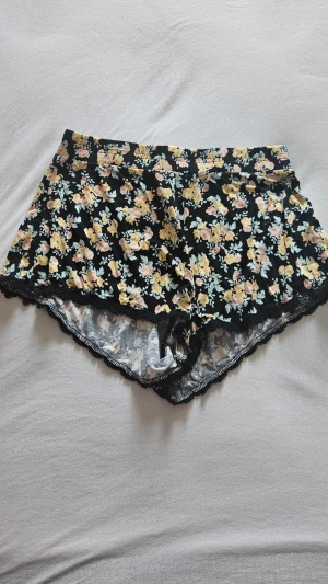 Blommiga shorts med spetskant - Snygga shorts med svart botten och blommigt mönster i gult, rosa och ljusblått. Shortsens kanter är dekorerade med svart spets och de har en bekväm resår i midjan. 