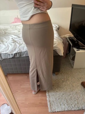 Beige långkjol med slits - Snygg beige långkjol med slits bak. Kjolen har en enkel och stilren design med låg midja och rak passform. Perfekt att matcha med crop top eller stickad tröja för en trendig look.