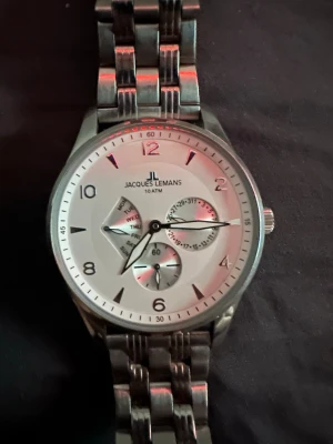 Jacques Lemans silverfärgad klocka - Stilren klocka från Jacques Lemans i silverfärgat metallarmband och boett. Urtavlan är vit med flera subdials för datum och veckodag. Klassisk design med tydliga siffror och markeringar, perfekt för dig som gillar en clean look. Den funkar helt o hållet men behöver ett nytt batteri.