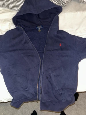 Marinblå hoodie från Polo Ralph Lauren - Snygg marinblå hoodie från Polo Ralph Lauren med dragkedja och huva. Klassisk röd logga broderad på bröstet och två fickor framtill. Tillverkad i mjuk bomullsblandning som känns skön mot huden. Perfekt för en avslappnad och stilren look. Passar mig som är 13 år 172cm och 55-60 kg