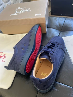 Blåa sneakers från Christian Louboutin - Snygga sneakers i mörkblå mocka från Christian Louboutin med ikonisk röd sula och diskreta detaljer. Skorna har klassisk låg profil, rund tå och matchande blå snörning. Perfekta för dig som vill sticka ut med exklusiv stil och lyxiga material. Storlek 42 och 44 finns. HELT NYA ALDRIG ANVÄNDa 10/10 skick. Alla tillbehör medföljer. 