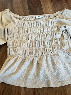 Beige offshoulder blus från JDY - Snygg beige offshoulder blus från JDY i storlek M. Blusen har smockad överdel och lätt puffiga ärmar. Tillverkad i ett tunt, strukturerat tyg som ger en luftig känsla. Perfekt för en trendig och avslappnad look, använd 1 gång.