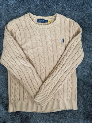 Beige Ralph Lauren Tröja - Beige Kabeldragen Ralph Lauren tröja i storlek M samt nyskick som endast är använd ett fåtal gånger. Perfekt till en sommarkväll, kom gärna med frågor vid funderingar eller prisförslag.