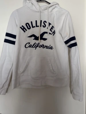 Vit hoodie från Hollister med tryck - Vit hoodie från Hollister med marinblå text och logga på bröstet samt två marinblå ränder på ärmarna. Klassisk känguruficka framtill, dragsko i huvan och mjukt material. Perfekt för en avslappnad och sportig stil.