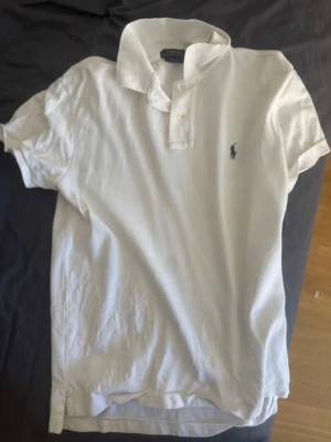 Vit pikétröja från Polo Ralph Lauren - Vit pikétröja från Polo Ralph Lauren med klassisk krage, korta ärmar och broderad logga på bröstet. Slim fit-modell i mjuk bomull, perfekt för en clean och stilren look.