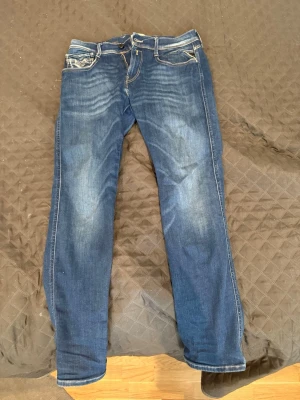 Replay jeans L30 W30 - Säljer ett par blå jeans med snygga kontrastsömmar och klassisk femficksdesign. Jeansen har en lätt tvättad look med slitningar på framsidan och bakfickor med dekorativa sömmar. Materialet är denim och passformen är rak.