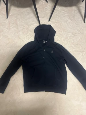 Mörkblå hoodie från Polo Ralph Lauren - Snygg mörkblå hoodie från Polo Ralph Lauren med dragkedja framtill och klassisk huva. Ikoniska broderade loggan på bröstet och två fickor framtill. Tillverkad i mjukt bomullsmaterial som är skönt att bära. Perfekt för en avslappnad och stilren look. Storlek L inte ens använd 