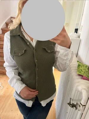Olivgrön ribbad väst med knappar - Snygg olivgrön väst med ribbad struktur och knäppning framtill. Västen har två bröstfickor med lock och en klassisk krage. Perfekt att styla över en skjorta för en trendig lager-på-lager-look.