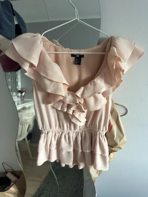 Ljusrosa volangtopp från H&M - Supersöt ljusrosa topp från H&M med volangdetaljer runt ringningen och midjan. Toppen är ärmlös och har en smickrande, lätt rynkad passform. 
