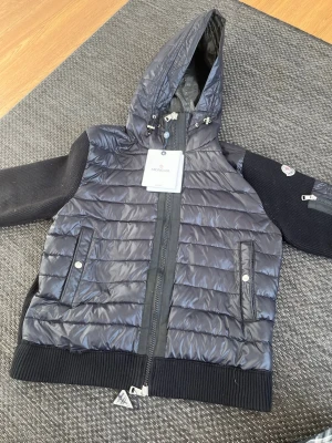 Svart pufferjacka från Moncler - Snygg svart pufferjacka från Moncler med glansig yta, stickade svarta ärmar och ribbade muddar. Jackan har huva med dragsko, två fickor med tryckknappar och dragkedja framtill. Perfekt för kalla vinterdagar och riktigt trendig look. Fraktar inte men kan mötas upp i Malmö 