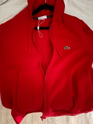 Röd zip hoodie från Lacoste - Snygg röd hoodie från Lacoste med dragkedja framtill och klassisk krokodil-logga på bröstet. Hoodien har huva med dragsko och två stora fickor framtill. Tillverkad i mjukt material som känns skönt mot huden. Perfekt för en avslappnad och sportig stil. Sälja pga den är för liten för mig nu