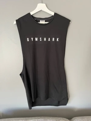 Svart Gymshark linne - Snyggt svart linne från Gymshark med vitt tryck framtill. Ärmlös modell med breda ärmhål, perfekt för gymmet eller träning. Tillverkat i mjukt och stretchigt material som ger bra rörelsefrihet.