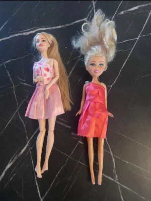 Två dockor i rosa klänningar - Säljer två dockor med långt hår, båda klädda i rosa klänningar. Den ena har blont hår och prickig klänning, den andra har ljusare hår uppsatt och en enkel rosa klänning. Perfekt för lek eller samling.