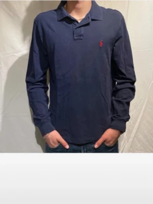 Marinblå långärmad piké från Polo Ralph Lauren - Klassisk marinblå långärmad pikétröja från Polo Ralph Lauren med röd broderad logga på bröstet. Skjortan har krage och knappar vid halsen. Tillverkad i mjuk bomull för en bekväm känsla och stilren look.