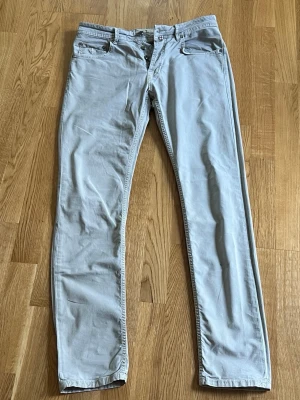 Ljusgrå chinos från Morris - Snygga ljusgrå chinos från Morris med klassisk rak passform och diskret broderad logga på myntfickan. Byxorna har fem fickor, bälteshällor och är tillverkade i mjukt bomullstyg med lite stretch för extra komfort.