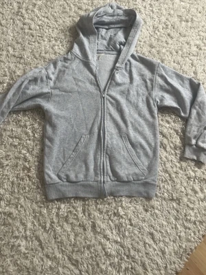 Grå zip hoodie från H&M - Klassisk grå hoodie från H&M med dragkedja framtill och två fickor. Tröjan har huva och ribbade muddar vid ärmslut och nederkant. Tillverkad i mjukt bomullsmaterial som är skönt att ha på sig. 