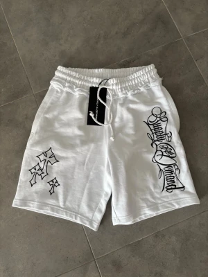 Vita shorts med broderi och snörning - Snygga vita shorts med svart broderad text och korsdetaljer. De har elastisk midja med snörning och är tillverkade i ett mjukt bomullsmaterial. Perfekta för en chill och trendig look.