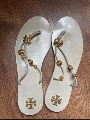 Tory burch Beige sandaler med gulddetaljer size US 8.5 - Stilrena beige sandaler med tunna remmar och dekorativa guldfärgade pärlor och logodetaljer. Platta sulor och öppen tå ger en avslappnad men trendig look. Perfekta för sommaren och lätta att matcha med olika outfits. Size US 8.5
