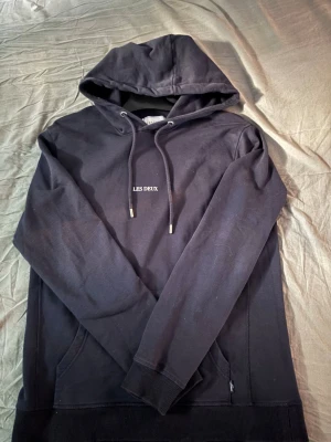 Svart hoodie från Les Deux - Säljer en svart hoodie från Les Deux med broderad vit logga på bröstet. Hoodien har dragsko i huvan, känguruficka och ribbade muddar vid ärmar och nederkant. Tillverkad i mjukt bomullstyg som är skönt att bära. Priset är förhandlingsbart.