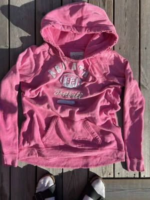 Rosa hoodie med tryck och huva - Säljer en rosa hoodie med huva och magficka. På bröstet finns ett broderat tryck med texten 'AB ATH 92 Football' och en amerikansk fotboll. Hoodien har långa ärmar och dragsko i huvan. Perfekt för en avslappnad och sportig stil.
