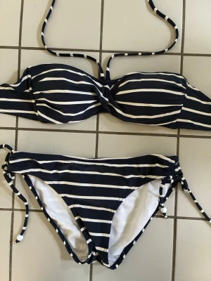 Randig blåvit bikini med knyt - Snygg bikini med marinblå och vita ränder. Överdelen har bandeau-modell med avtagbara axelband och twistad front. Underdelen har knyt i sidorna för justerbar passform. Perfekt för strandhäng eller pooldagar☀️Den är helt oanvänd, men saknar prislapp!