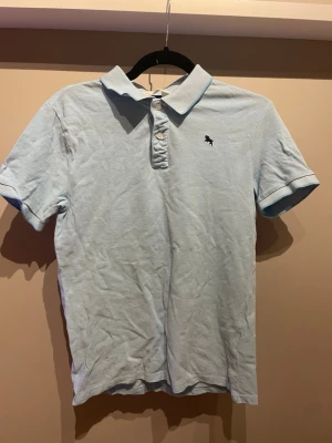 Ljusblå pikétröja från H&M - Säljer en ljusblå pikétröja från H&M med klassisk krage och knappar framtill. Tröjan har korta ärmar och en liten broderad logga på bröstet. Perfekt för en avslappnad och stilren look.