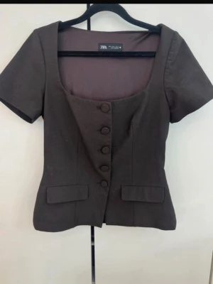 Brun kortärmad blus från Zara - Stilren brun blus från Zara med korta ärmar och markerad midja. Djup fyrkantig ringning och klädda knappar framtill ger en elegant look. Perfekt att matcha med både jeans och kjol.