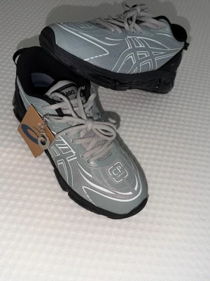 Grå sneakers från Asics x CP Company - Snygga sneakers i samarbete mellan Asics och CP Company. Skorna är grå med vita och svarta detaljer, har reflekterande mönster och en chunky svart sula. Ovandelen är i mesh och syntetmaterial, med klassiska Asics-loggor på sidorna och CP Company-märke på tån.