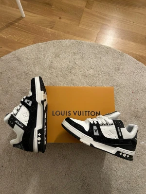 Louis Vuitton svartvita sneakers - Snygga svartvita Louis Vuitton sneakers med klassisk LV-logga på plösen och sidan. Skorna har vita snören, svarta detaljer och monogrammönster i material som känns lyxigt och slitstarkt. Perfekta för dig som vill sticka ut med exklusiv streetstyle.