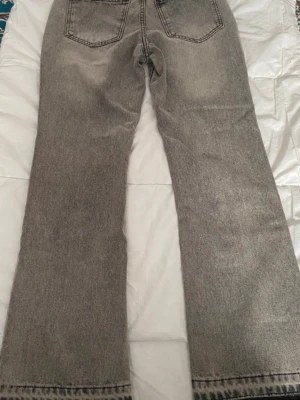 Grå bootcut jeans med rå kant - Säljer ett par grå bootcut jeans med snygg tvätt och rå, fransad kant längst ner. Jeansen har klassiska fem fickor och raka ben som går ut lite nedtill. Perfekta för en avslappnad och trendig look.