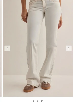 Vita bootcut jeans med kontrastsömmar - Säljer ett par vita bootcut jeans med snygga kontrastsömmar och klassiska fem fickor. Jeansen har låg midja och utsvängda ben, samt dekorativa sömmar på bakfickorna. Perfekta för en trendig och avslappnad look.
