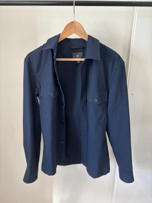 Mörkblå overshirt - Stilren mörkblå overshirt från H&M med klassisk krage och två bröstfickor med knappar. Skjortan har långa ärmar och knäppning framtill. Perfekt för en clean och enkel look.