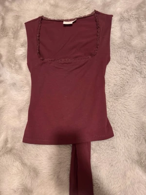 Vinröd topp med knyt - Snygg vinröd topp i mjukt material med fyrkantig ringning och spetsdetalj runt halsen. Toppen är ärmlös och har en unik omlottdesign med knyt i ryggen som ger en cool och trendig look.
