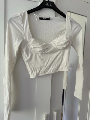 Vit croppad topp från Bik Bok - Säljer en vit croppad topp från Bik Bok med långa ärmar och markerade kupor. Toppen har en snygg, figurnära passform och djup urringning som ger en trendig look. Perfekt att styla med höga jeans eller kjol.