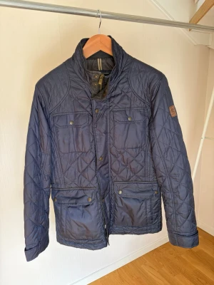 Marinblå quiltad jacka med fickor - Snygg marinblå quiltad jacka med fyra framfickor och hög krage. Jackan har tryckknappar och dragkedja framtill samt dekorativa sömmar i rutmönster. Perfekt för dig som vill ha en stilren och funktionell jacka i lätt material.