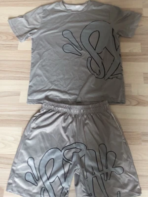 Syna world set - Säljer ett beige set med t-shirt och shorts i syntetmaterial. Båda delarna har ett stort svart grafiskt tryck framtill och baktill. T-shirten är kortärmad och shortsen har resår i midjan för skön passform. Perfekt för dig som gillar streetwear vibes.