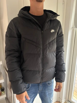 Svart pufferjacka från Nike med huva - Nypris 3500, Snygg svart pufferjacka från Nike med huva och dragkedja framtill. Jackan har två sidofickor med dragkedja och en vit Nike-logga på bröstet. Perfekt för kalla vinterdagar och ger en sportig look.